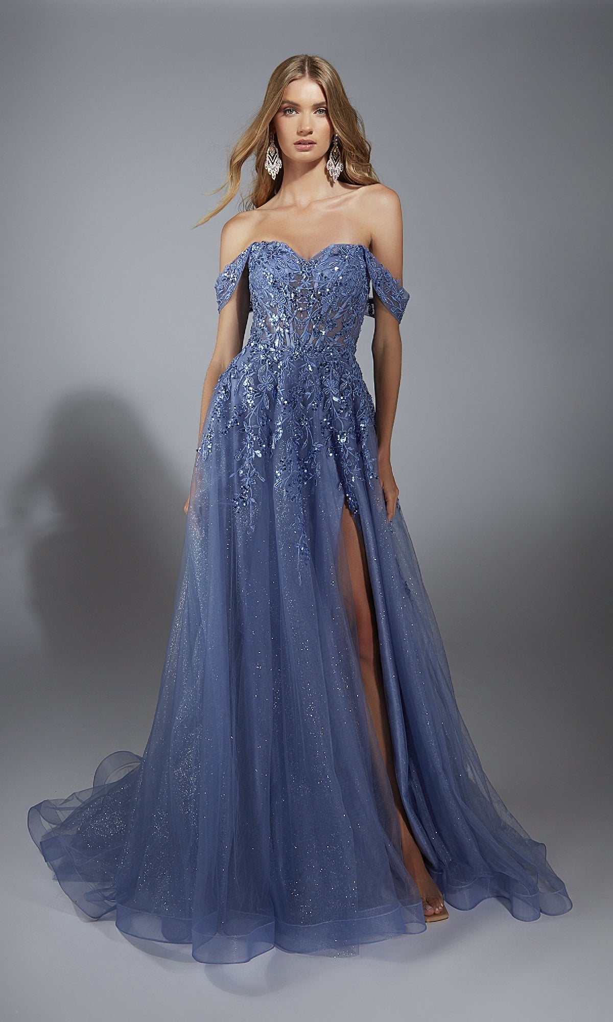 A woman wears the Aerie Couture 284 long prom dress, a blue gown with an off-the-shoulder corset bodice, floral embellishments, glitter tulle, a thigh-high slit, and an A-line skirt as she stands against a gray background.