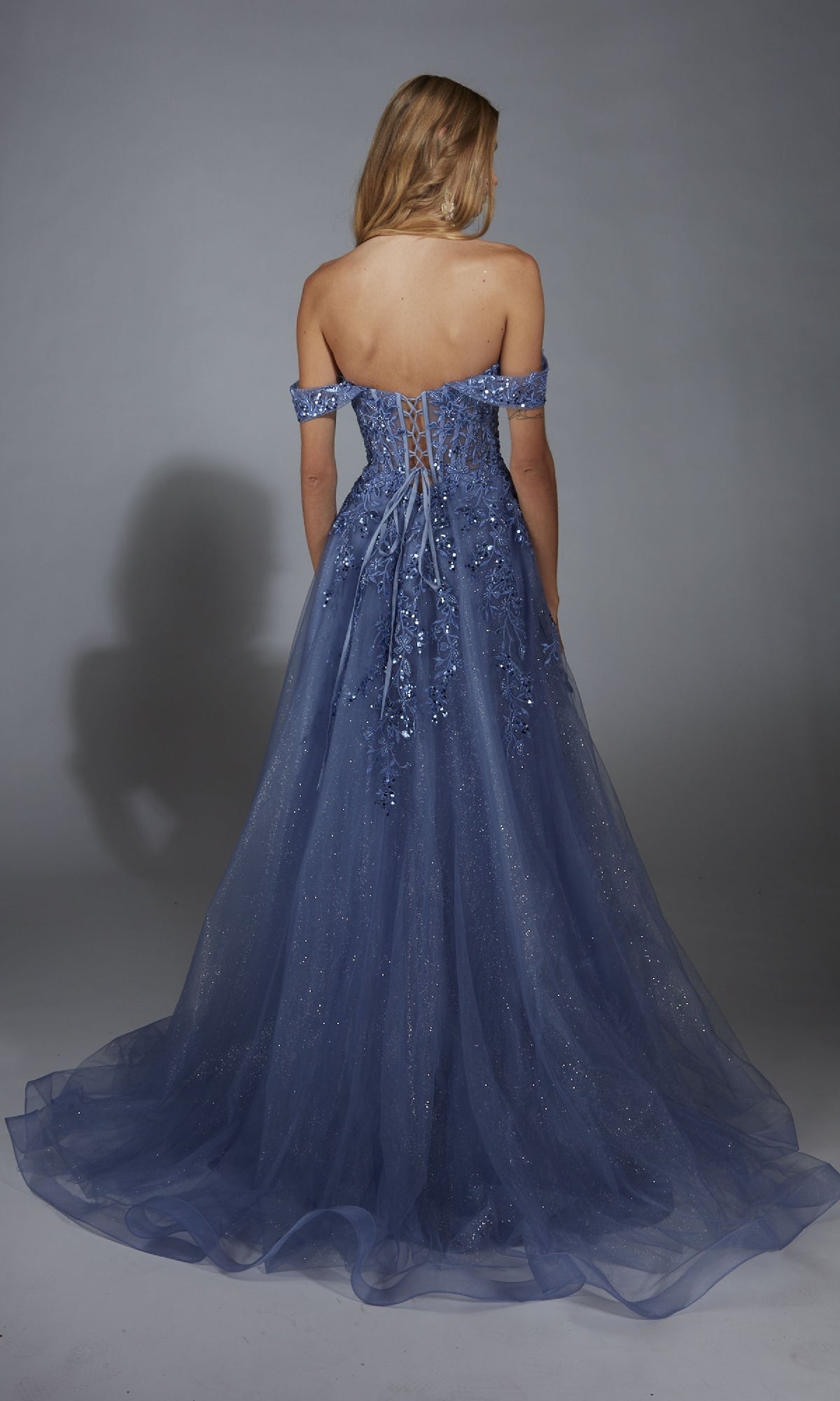 A woman stands facing away in front of a plain grey backdrop, wearing the Aerie Couture 284—an off-the-shoulder, floor-length glitter tulle prom dress featuring floral embroidery and an elegant a-line skirt.