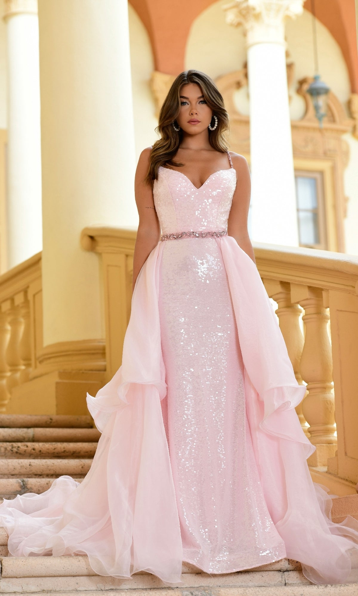 A woman stands on stairs wearing the Long Formal Dress 28560 by Ava Presley, a light pink sequin gown with a sweetheart neckline, beaded waist, and chiffon overskirt. Elegant architectural columns rise in the background.