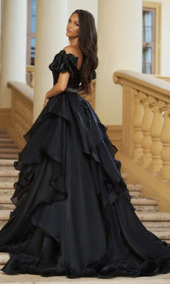 A woman stands on a grand staircase, looking back over her shoulder in the Ava Presley 28571 Puff-Sleeve Long Prom Ball Gown—a stunning black dress with elegant ruffled details.