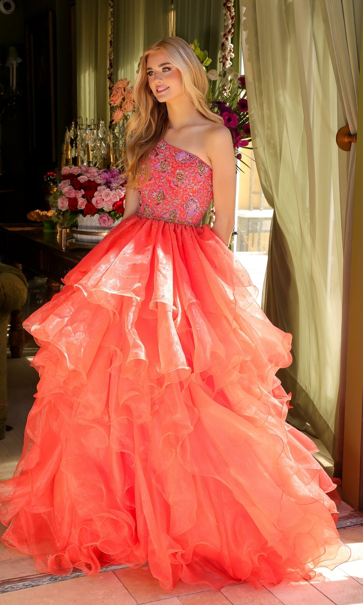 A woman stands indoors in the Ava Presley 28575 One-Shoulder Prom Ball Gown with a tiered, ruffled coral skirt and embellished bodice, as flowers and green curtains decorate the background.