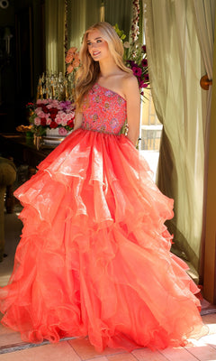 A woman stands indoors in the Ava Presley 28575 One-Shoulder Prom Ball Gown with a tiered, ruffled coral skirt and embellished bodice, as flowers and green curtains decorate the background.