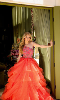 A woman in the Ava Presley 28575 one-shoulder prom ball gown with a bright coral, tiered skirt and floral embroidered bodice stands in a doorway, holding the door open with one hand.