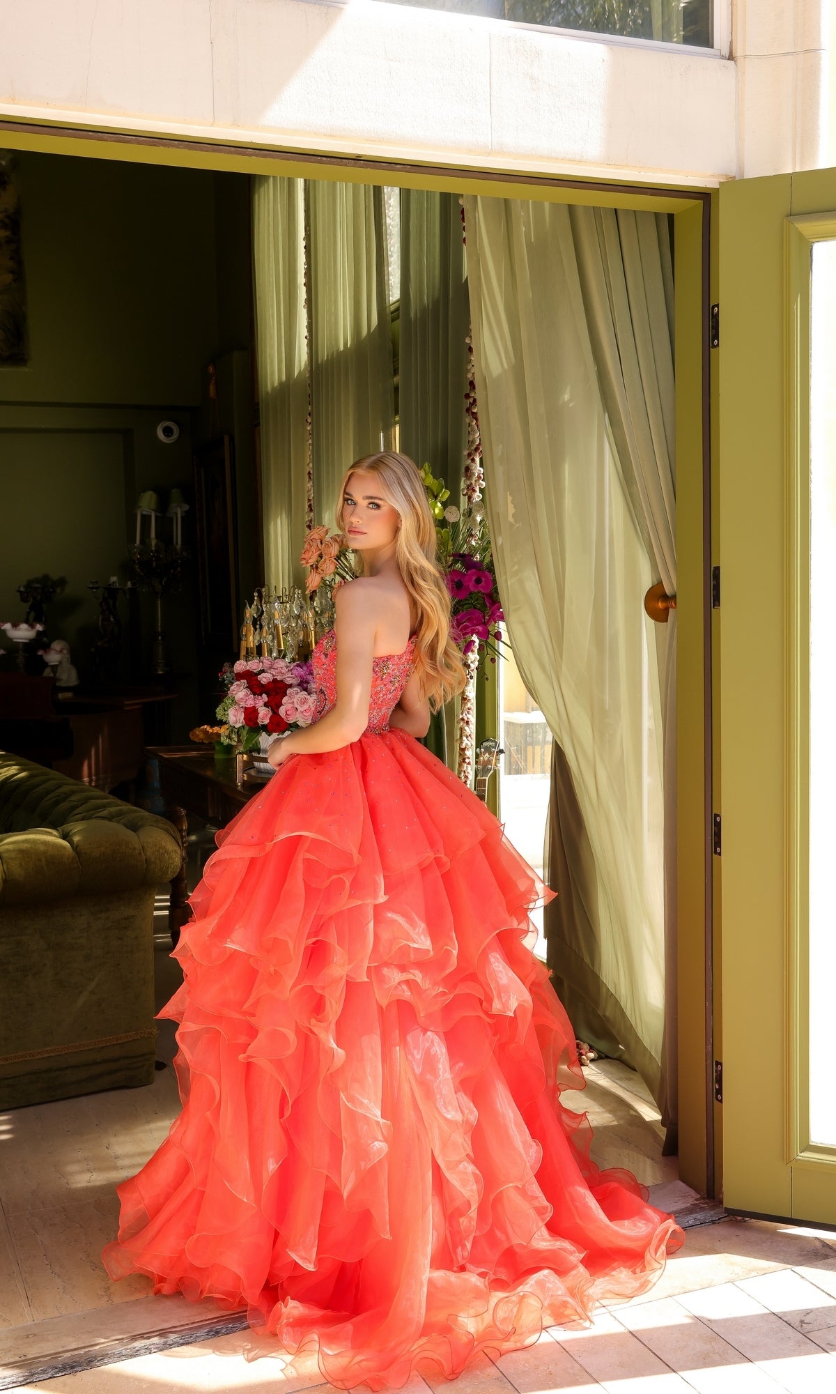 A woman in a bright coral Ava Presley 28575 one-shoulder prom ball gown stands in a doorway with green walls and floral decor, looking back over her shoulder.