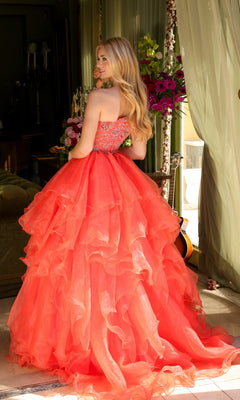 A woman with long blonde hair wears the Ava Presley 28575 One-Shoulder Prom Ball Gown in coral, featuring a voluminous ruffled skirt, as she stands indoors near curtains and flowers.