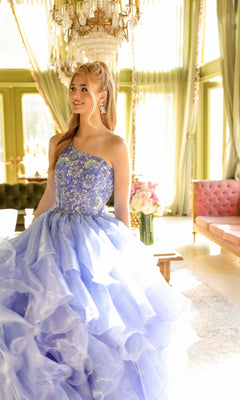 A woman stands in a well-lit, elegant room wearing the Ava Presley 28575 one-shoulder prom ball gown, perfect for those seeking a sophisticated and graceful long tiered dress for prom.