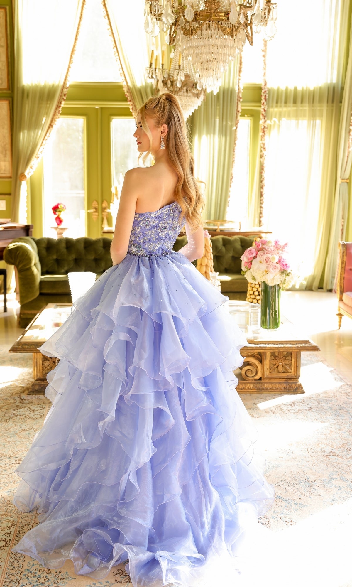 A woman in the One-Shoulder Prom Ball Gown: Ava Presley 28575 stands in an elegant, sunlit room with green walls, chandeliers, and large windows.