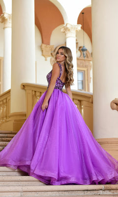 A woman wearing the Long Formal Dress 28579 by Ava Presley, featuring an embroidered bodice and v-neckline, stands on a staircase in an elegant architectural setting, looking back over her shoulder.