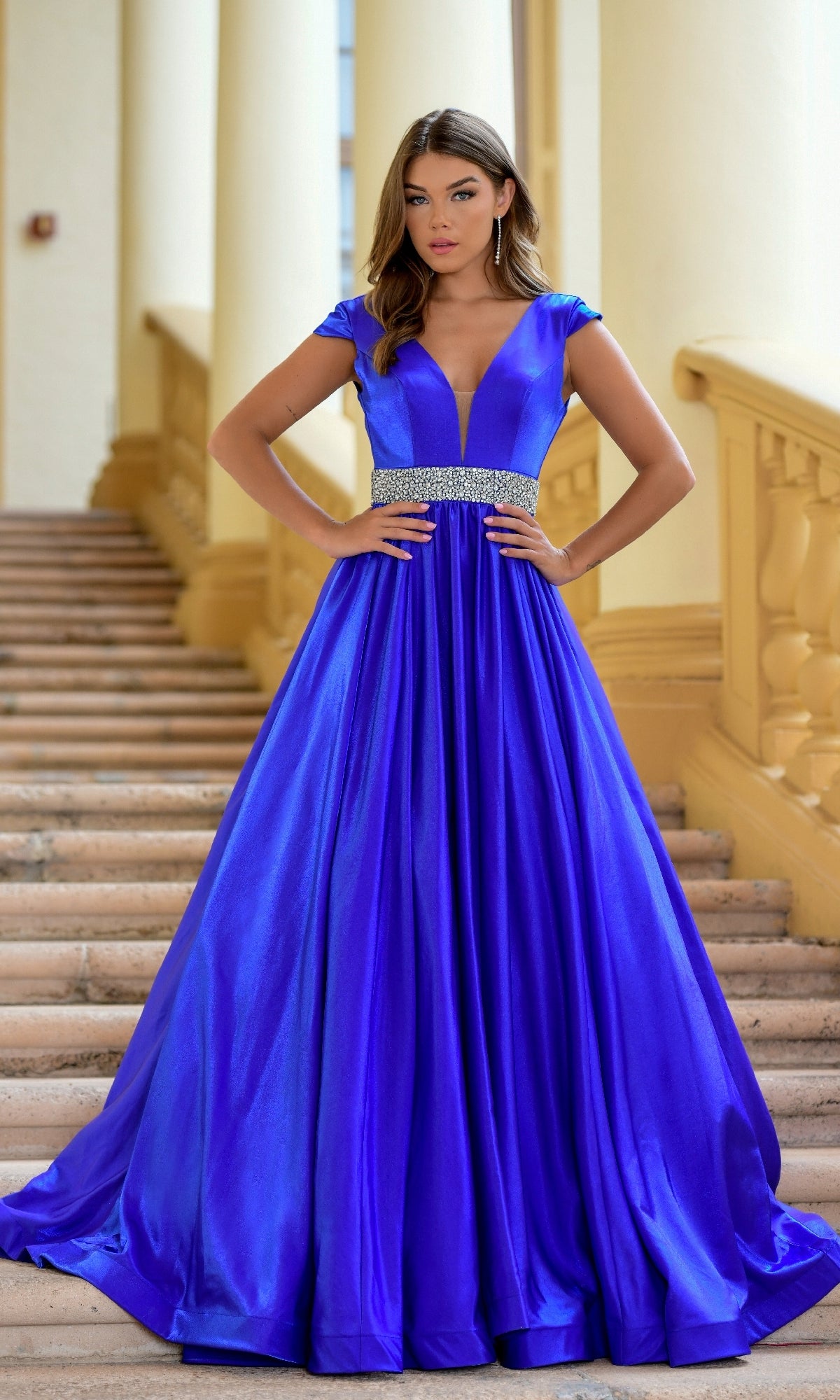 A woman stands on stairs wearing the Long Formal Dress 28580 by Ava Presley, featuring cap sleeves, a deep V-neckline, a jeweled waistband, and royal blue color.
