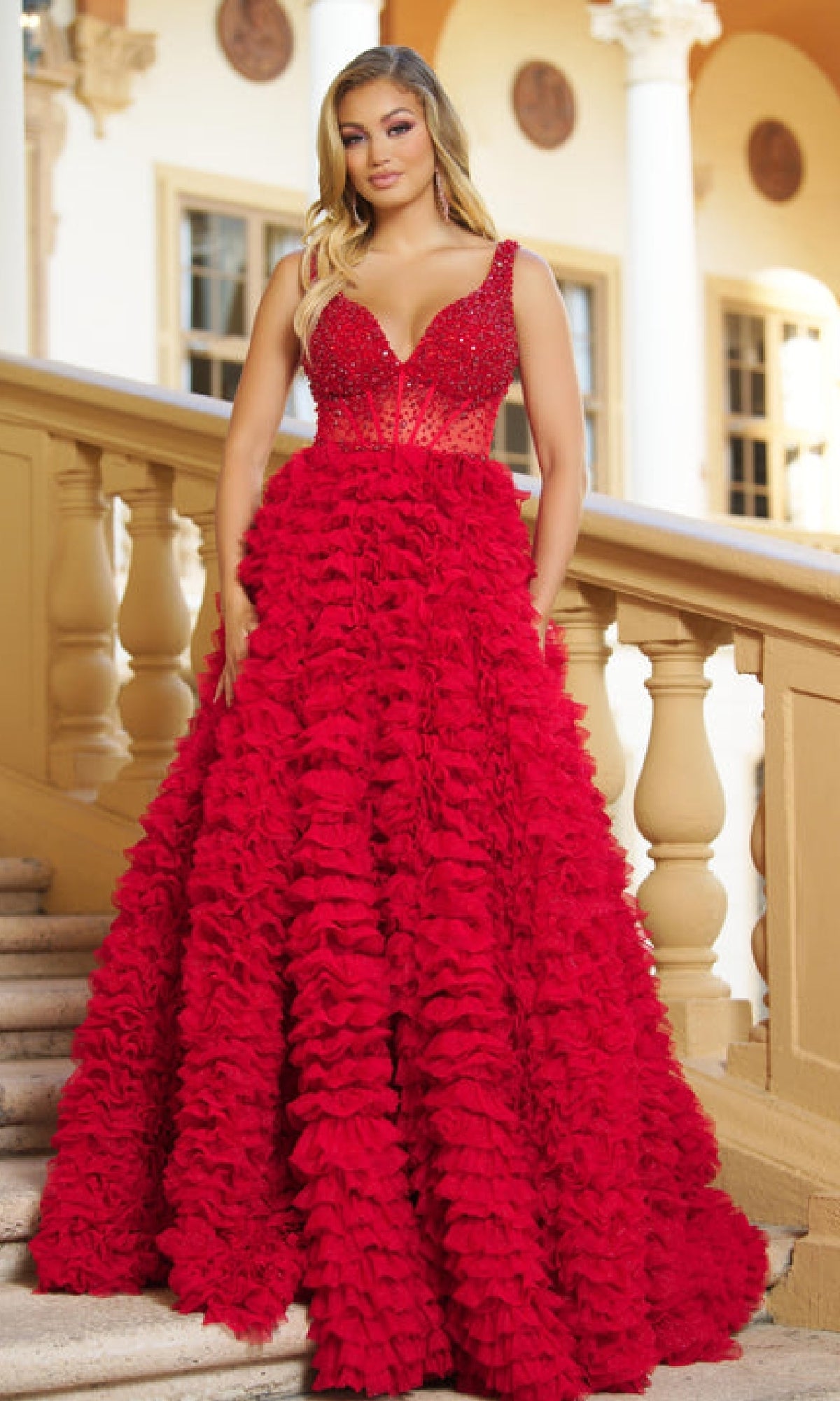 A woman stands on a staircase in the Ava Presley 28584 long ruffled prom ball gown, featuring a fitted beaded corset bodice and a voluminous tiered skirt. Columns and windows appear in the background.