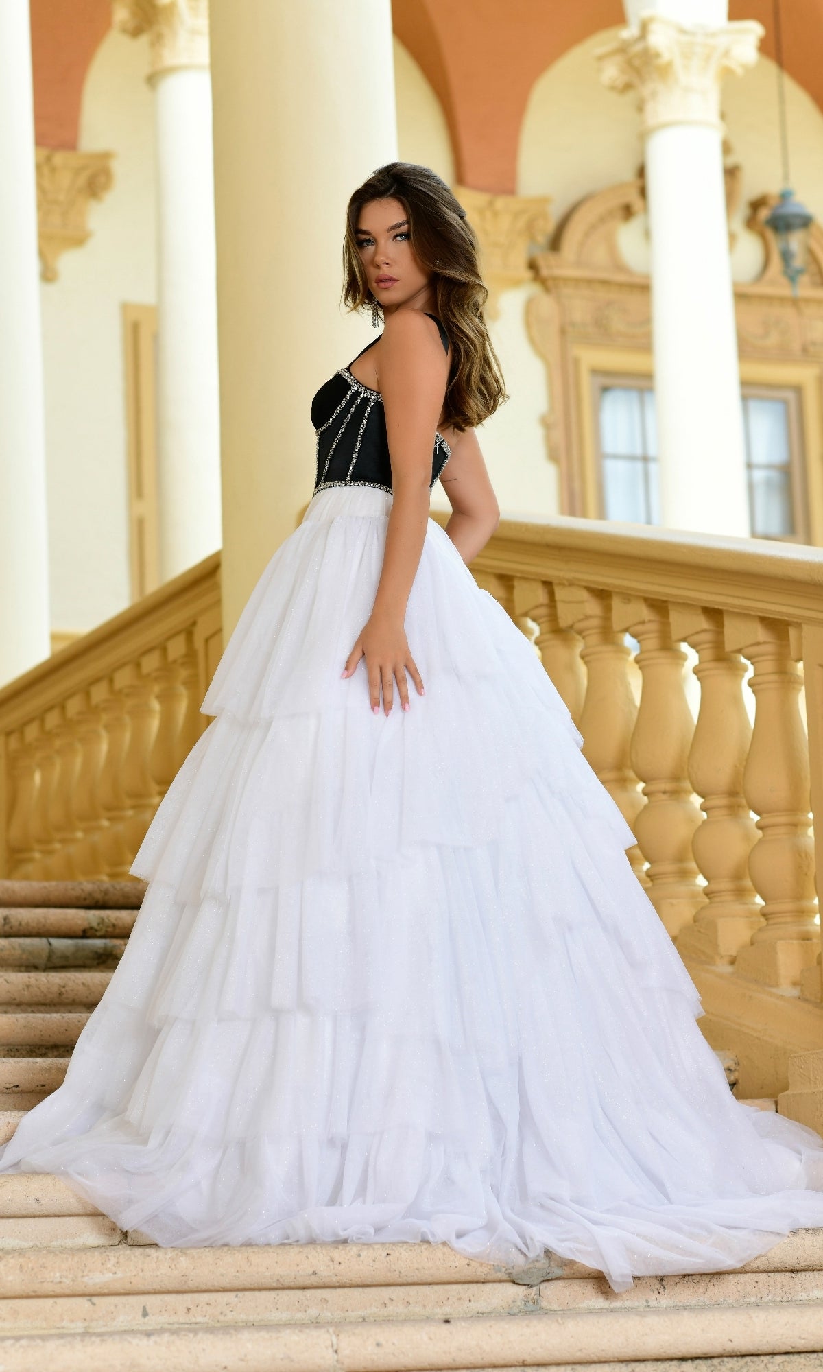 A woman stands on an outdoor staircase in the Long Formal Dress 28592 by Ava Presley, a black and white ball gown with an elegant A-line skirt, framed by columns and ornate architecture in the background.