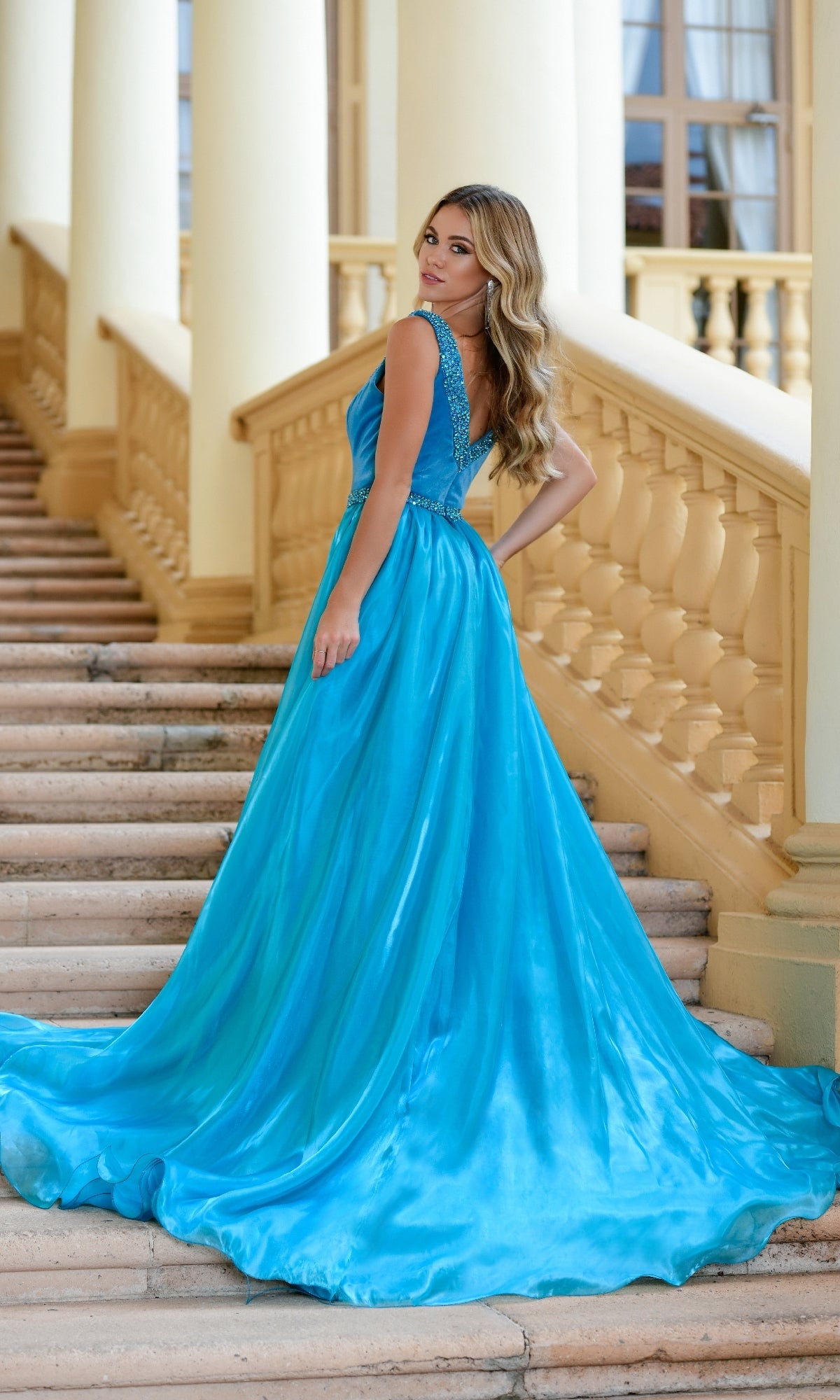 A woman wearing the Long Formal Dress 28599 by Ava Presley, a bright blue v-neck gown with a thigh-high slit, stands on a staircase and looks over her shoulder; behind her is a building with columns and large windows.