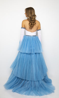 A woman faces away, wearing the Strapless High-Low Prom Gown: Ava Presley 28601 in blue tiered tulle with white gloves, set against a plain white background.