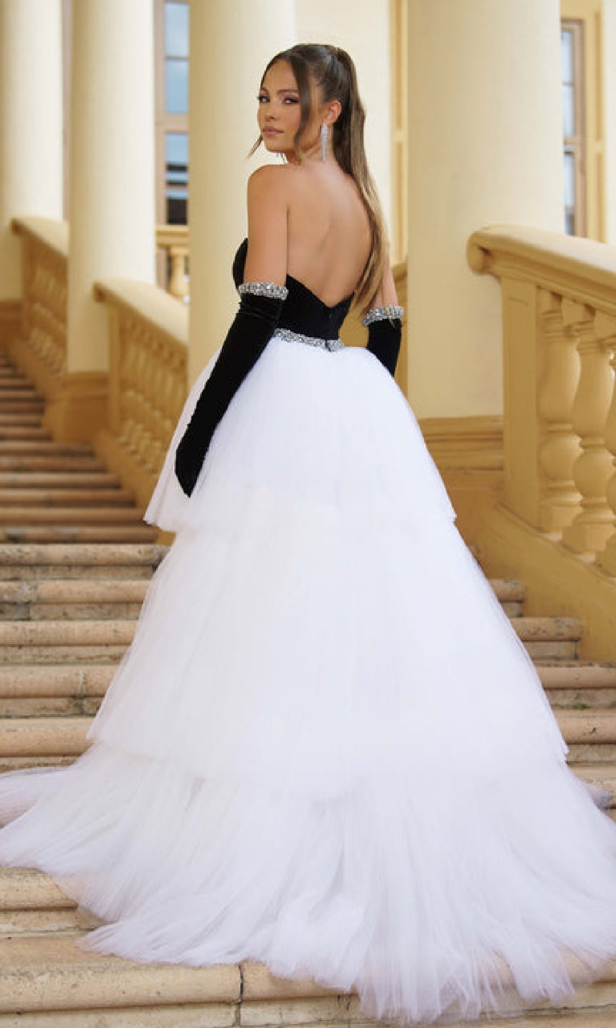 A woman stands on steps, elegantly looking over her shoulder in the Strapless High-Low Prom Gown: Ava Presley 28601, styled with black gloves and a tiered tulle skirt.