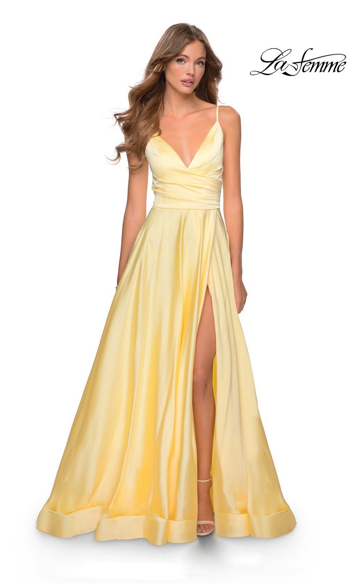 A woman wears the La Femme V-Neck Long A-Line Prom Dress 28607 in pale yellow with a deep V-neckline, thin straps, and a high slit, standing against a white background. The 