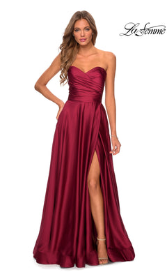 A woman poses against a white background in the La Femme Strapless Long A-Line Prom Dress 28608, a deep red satin gown featuring a sweetheart neckline, floor-length hem, and thigh-high slit.