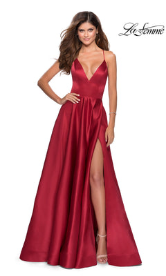 A woman poses elegantly in the La Femme Strappy-Back Long A-Line Prom Dress 28628, a deep red satin gown with a V-neckline, thigh-high slit, and spaghetti straps, against a plain white background.