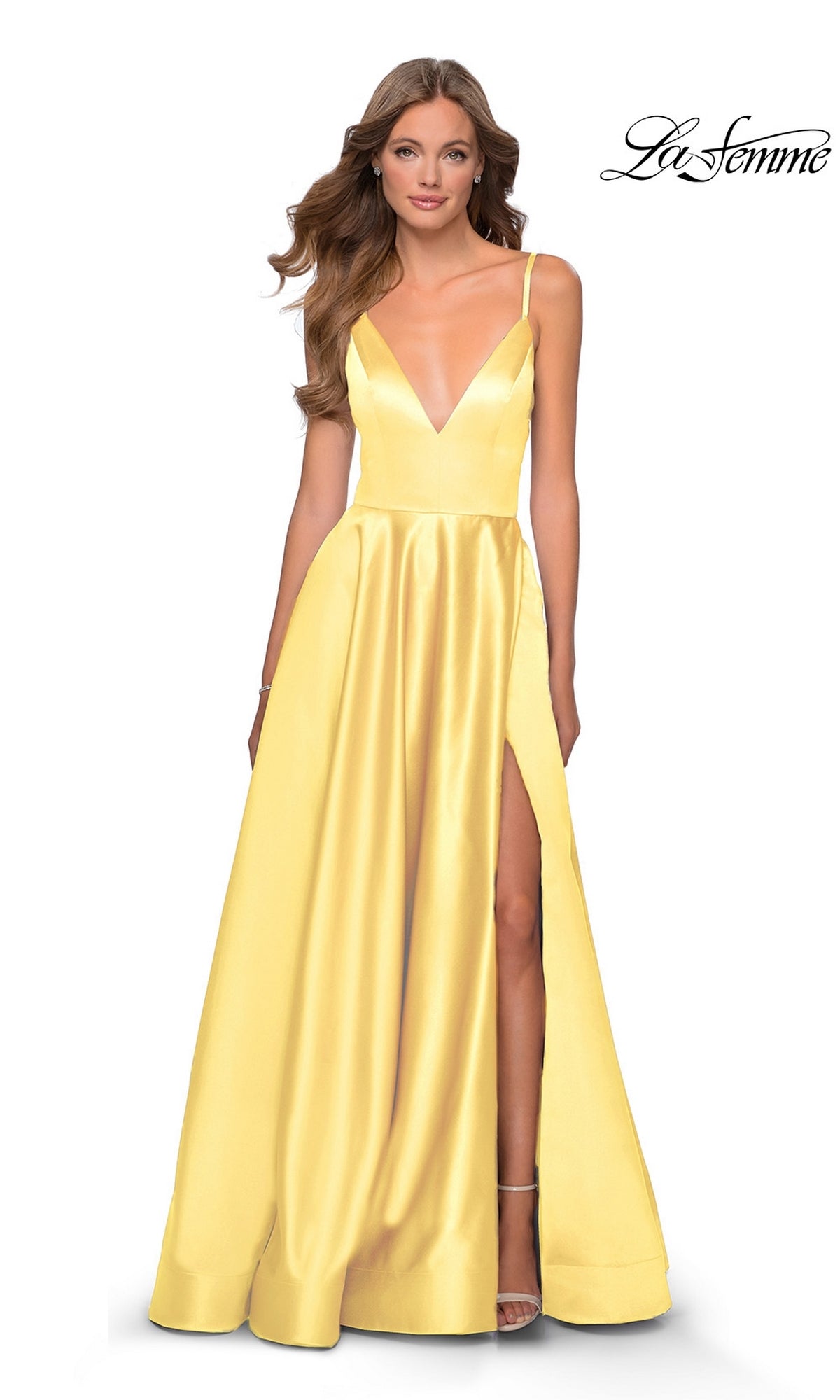 A woman wears the La Femme Strappy-Back Long A-Line Prom Dress 28628, a floor-length yellow satin v-neck gown with thin straps and a high slit, standing against a white background. The 