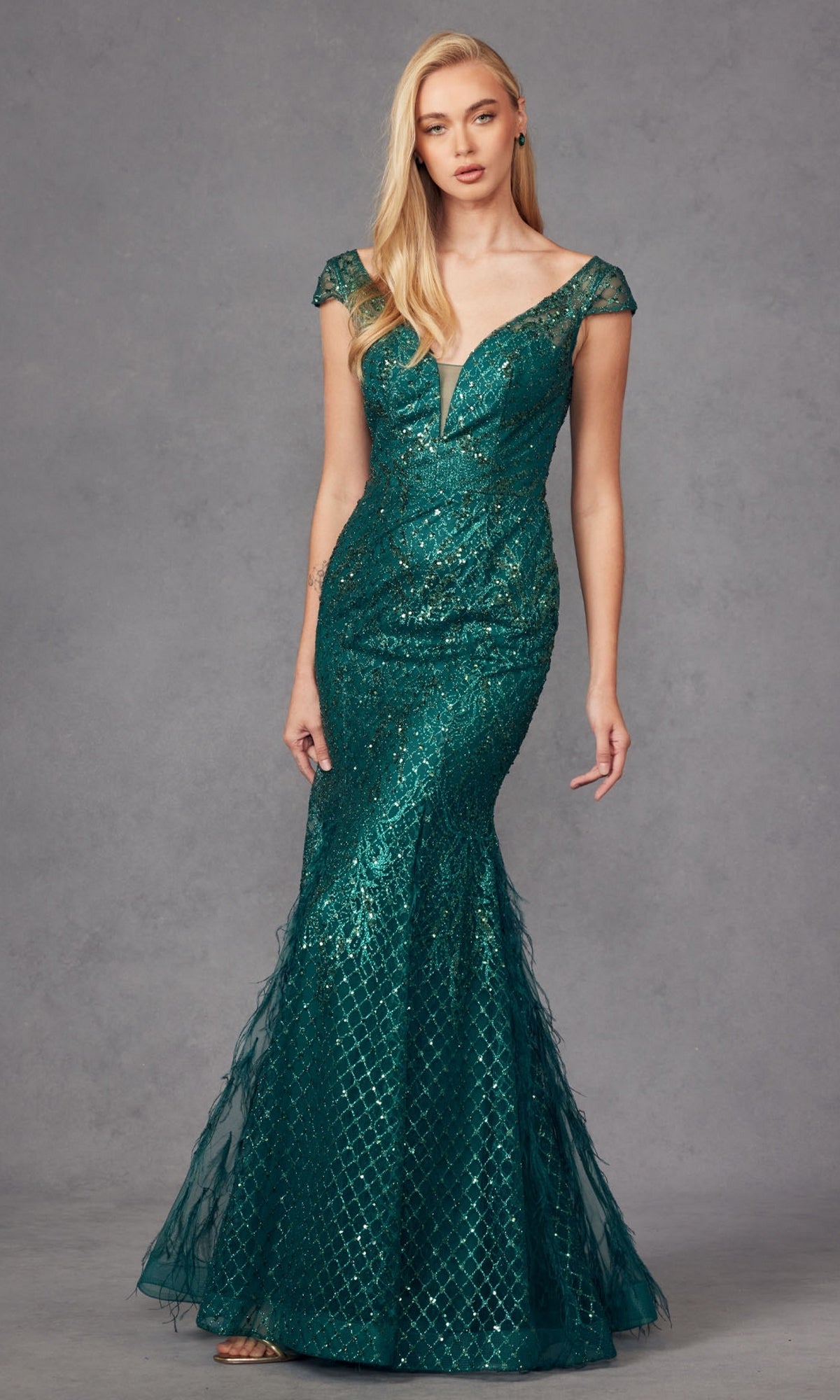 A woman stands against a gray studio backdrop wearing the Juliet 286 Feathered Cap-Sleeve Long Prom Dress—a fitted mermaid gown with feathered cap sleeves and a flared hem.
