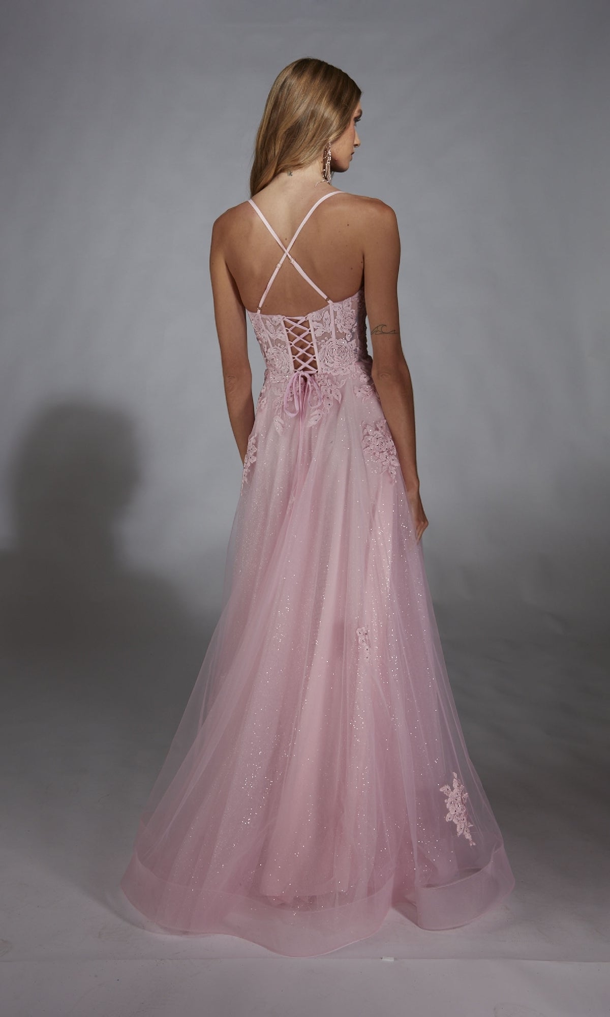 A woman faces away, wearing the Glitter Tulle Prom Ball Gown: Aerie Couture 286—a long pink corset-back dress with sheer overlay and floral accents—against a plain gray background.