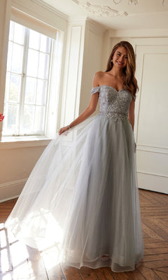 A woman stands in a sunlit room wearing the Juliet 287 Long Sweetheart Prom Ball Gown, featuring floral embroidery, a beaded corset bodice, and a flowing light gray tulle skirt that she gracefully holds to one side.