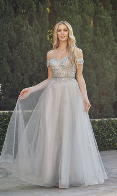 A woman stands outdoors before lush green hedges, wearing the Juliet 287 Long Sweetheart Prom Ball Gown—a silver off-the-shoulder dress with a beaded corset bodice and flowing tulle skirt.