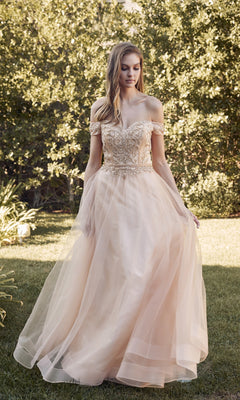 A woman stands outdoors in a garden wearing the Long Sweetheart Prom Ball Gown: Juliet 287, featuring an off-the-shoulder style with a light champagne tulle skirt and an intricately beaded corset bodice.