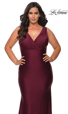 A woman wears the Long Plus-Size Prom Dress: La Femme 29016, a floor-length, deep burgundy jersey gown with a V-neckline and a faux wrap-style bodice.
