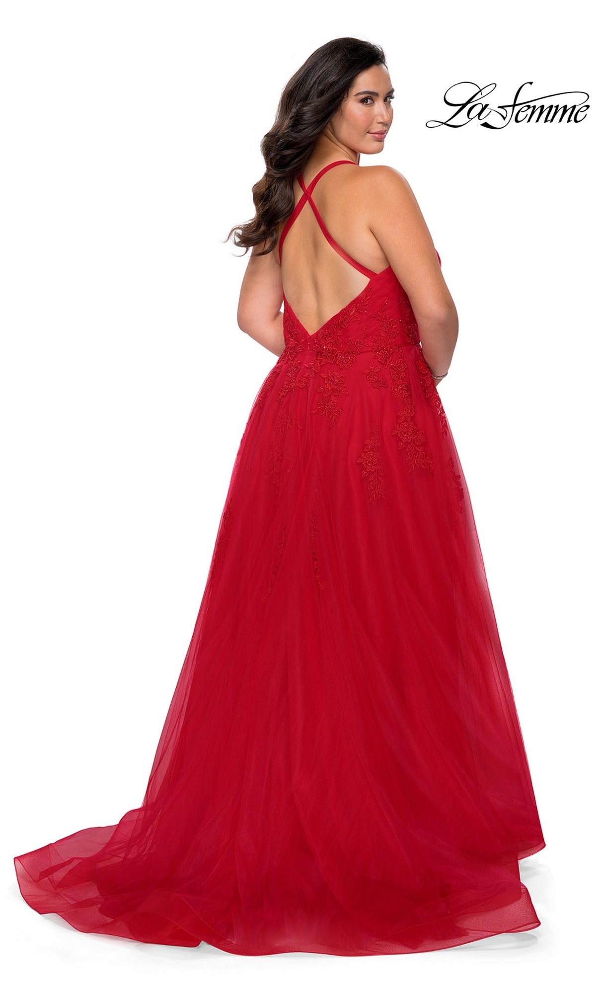 A woman models the La Femme Long Prom Dress 29021, a formal evening gown in vibrant red featuring delicate lace details, crisscross straps, and a low back. She looks over her shoulder as the flowing tulle skirt adds elegance. The “La Femme” logo appears in the top right corner.