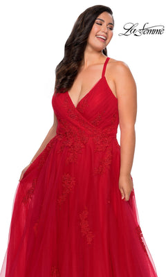 A woman smiles and holds the La Femme Long Prom Dress 29021—a red, sleeveless, floor-length formal evening gown with lace detailing—posed against a plain white background with the “La Femme” logo visible.