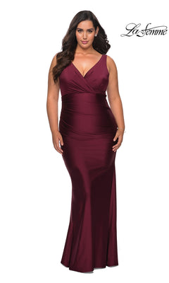 A woman wears a dark berry-colored Long Plus-Size Prom Dress: La Femme 29028, featuring a floor-length formal design with a V-neck and a ruched bodice.