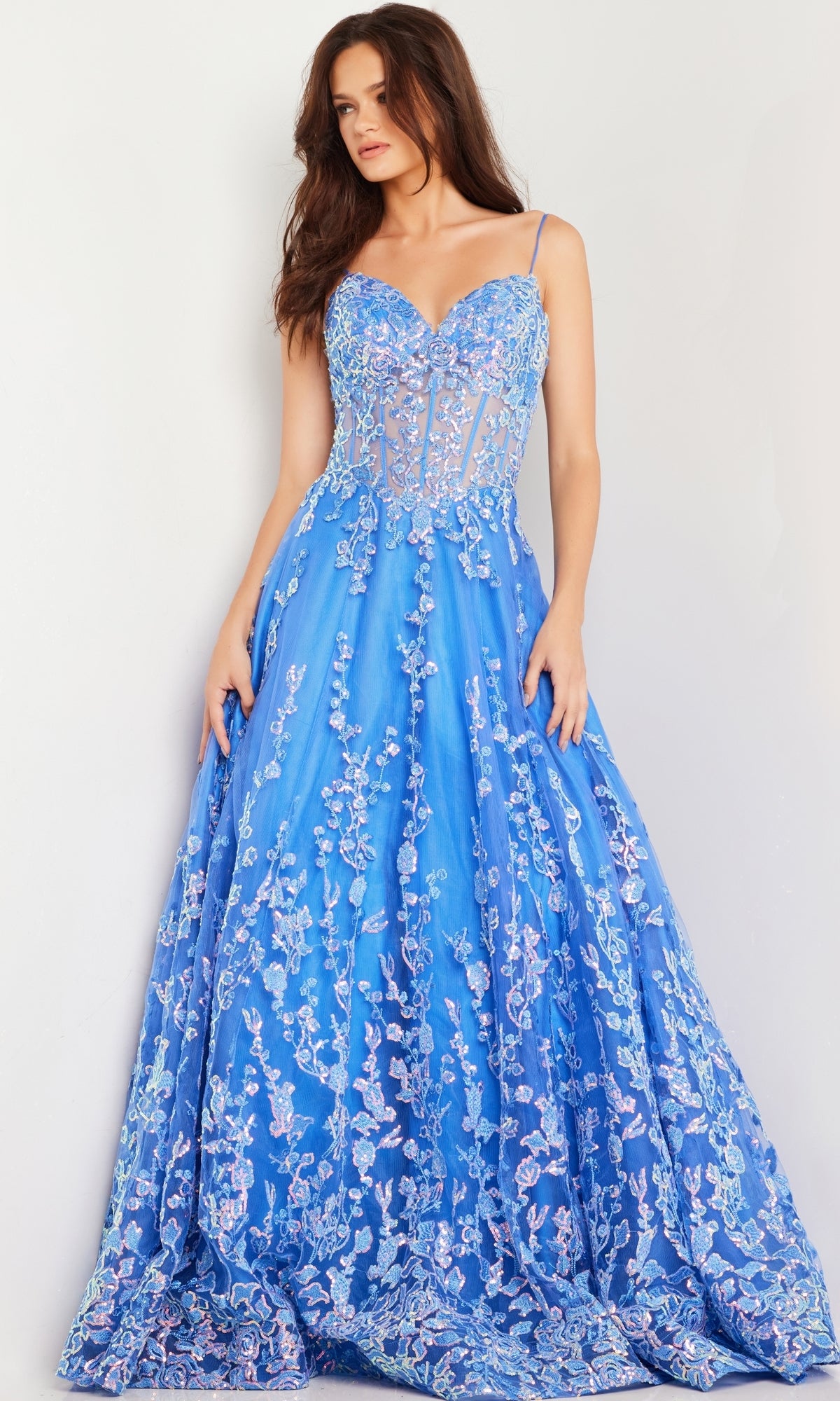 A woman stands against a light background wearing Jovani Long Prom Dress 29072—a floor-length turquoise blue gown with a sheer corset bodice, spaghetti straps, and floral sequin embroidery.