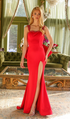 A woman wears the Long Prom Dress 29121 by Ava Presley, a fitted red gown with a high slit, standing indoors on a patterned rug before tall windows with green curtains and a green velvet sofa.