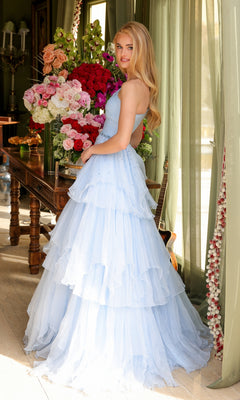 A woman in the Ava Presley 29131 One-Shoulder Tiered Prom Gown stands indoors by a table with large floral arrangements and a guitar leaning against the wall.