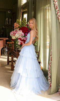 A woman wearing the Ava Presley 29131 One-Shoulder Tiered Prom Gown in light blue stands beside a table with large flower bouquets in a sunlit room with green curtains.