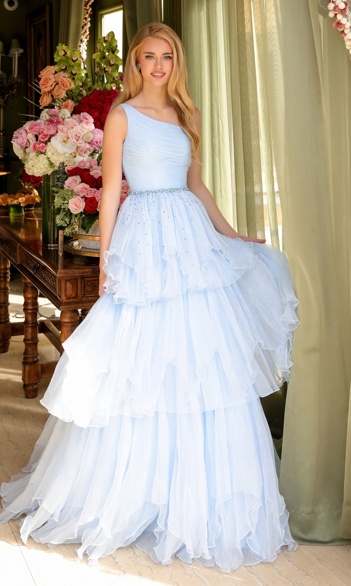 A woman in the Ava Presley 29131 One-Shoulder Tiered Prom Gown, light blue, stands indoors by a table with floral arrangements and sheer green curtains in the background.