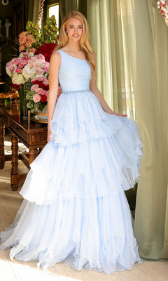 A woman in the Ava Presley 29131 One-Shoulder Tiered Prom Gown, light blue, stands indoors by a table with floral arrangements and sheer green curtains in the background.