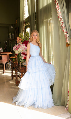 A woman in the Ava Presley 29131 One-Shoulder Tiered Prom Gown stands indoors by a window with sheer green curtains, beside a flower-adorned table.