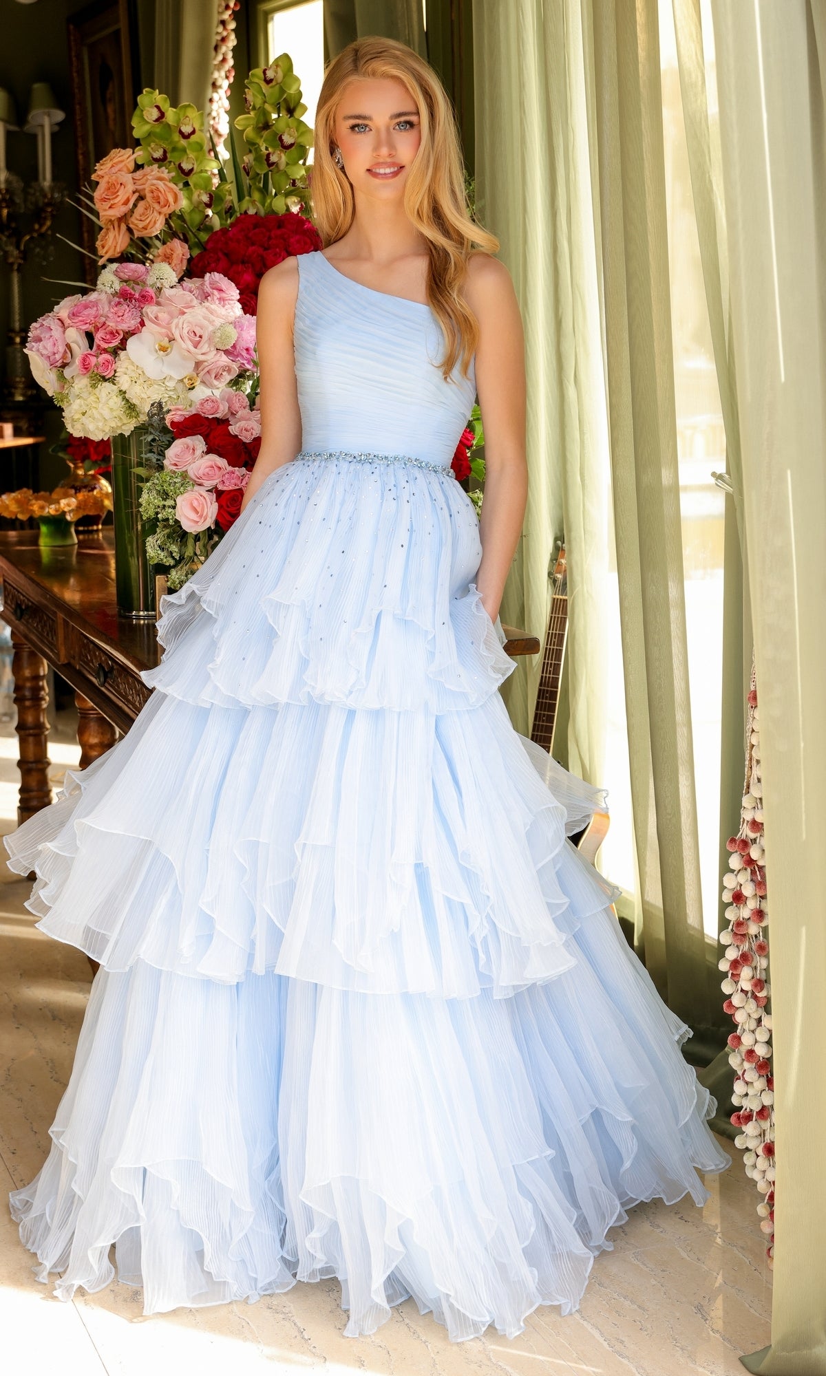 A woman in the Ava Presley 29131 One-Shoulder Tiered Prom Gown, light blue and floor-length, stands indoors by a window with green curtains and a large floral arrangement in the background.