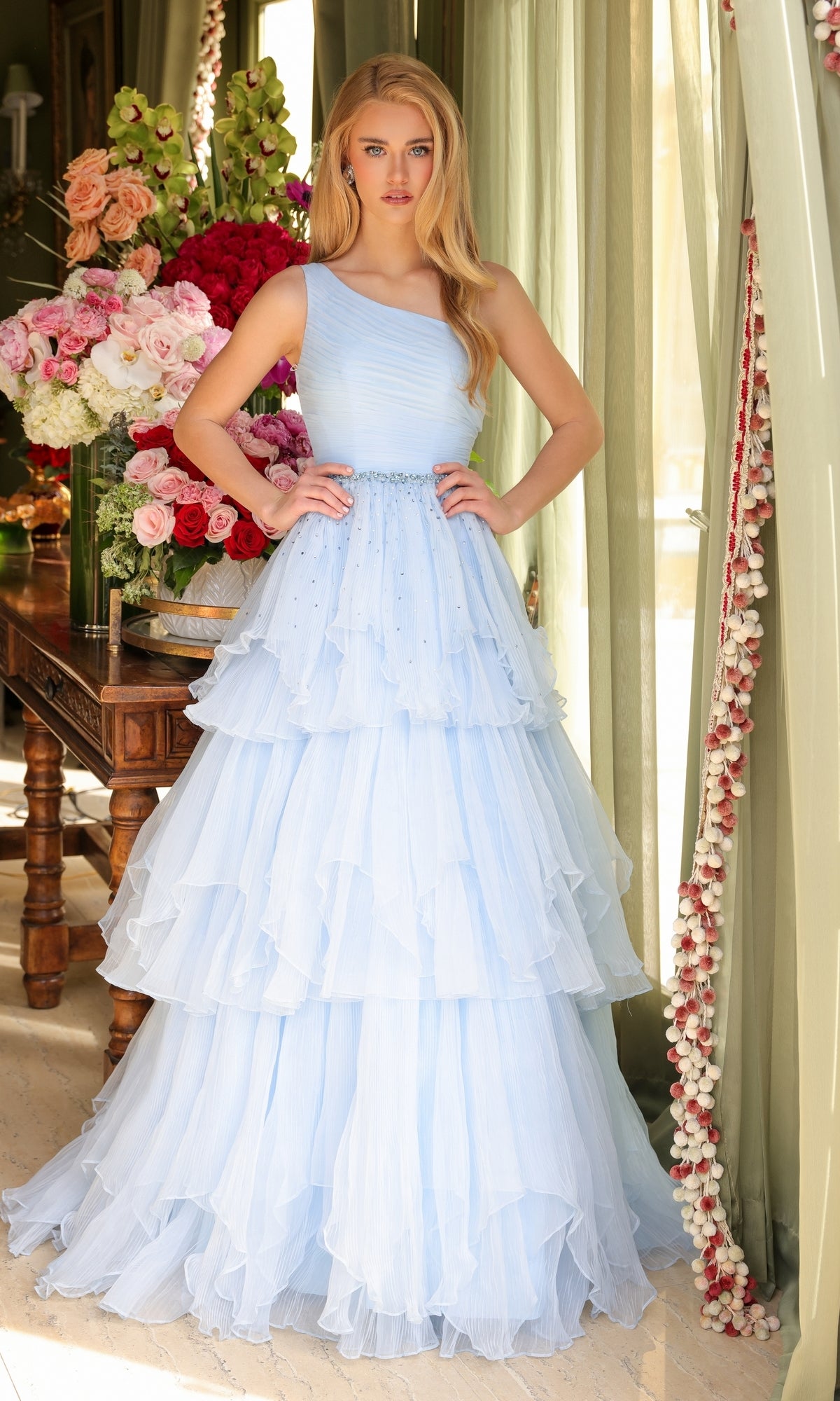 A woman stands indoors beside a table with large floral arrangements and green curtains, wearing the Ava Presley 29131 One-Shoulder Tiered Prom Gown in light blue with ruffled, tiered layers.