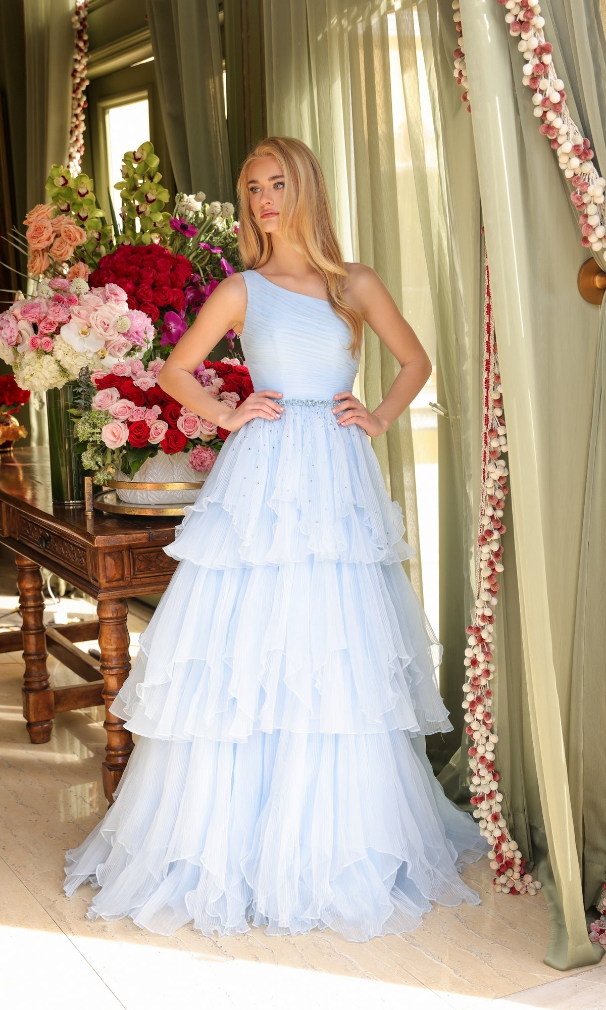 A woman in the Ava Presley 29131 one-shoulder tiered prom gown stands by a window with green curtains and a table adorned with large pink and red flower arrangements.