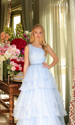 A woman stands indoors by a table of colorful flowers, wearing the Ava Presley 29131 One-Shoulder Tiered Prom Gown in light blue as sunlight streams through sheer green curtains behind her.