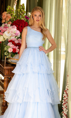 A woman stands indoors by a table with pink and white flowers, wearing the Ava Presley 29131 One-Shoulder Tiered Prom Gown in light blue.