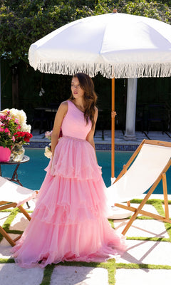 A woman in the Ava Presley 29131 One-Shoulder Tiered Prom Gown stands by a pool under a white fringed umbrella, with deck chairs and a flower arrangement nearby.