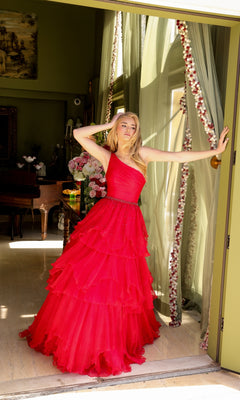 A woman in the Ava Presley 29131 one-shoulder tiered prom gown stands in a floral garland-adorned doorway, posing with one arm raised and the other touching the frame.