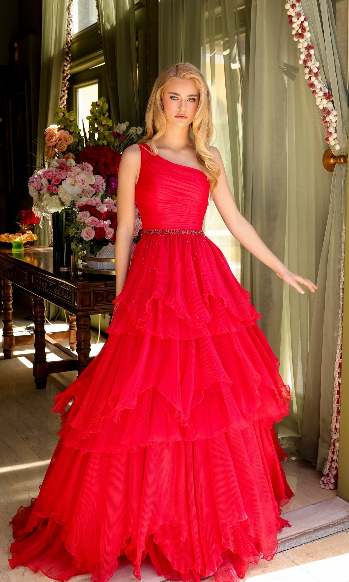 A woman stands indoors wearing the Ava Presley 29131 One-Shoulder Tiered Prom Gown, a vibrant red dress with beaded accents, framed by green curtains and a floral-adorned table.