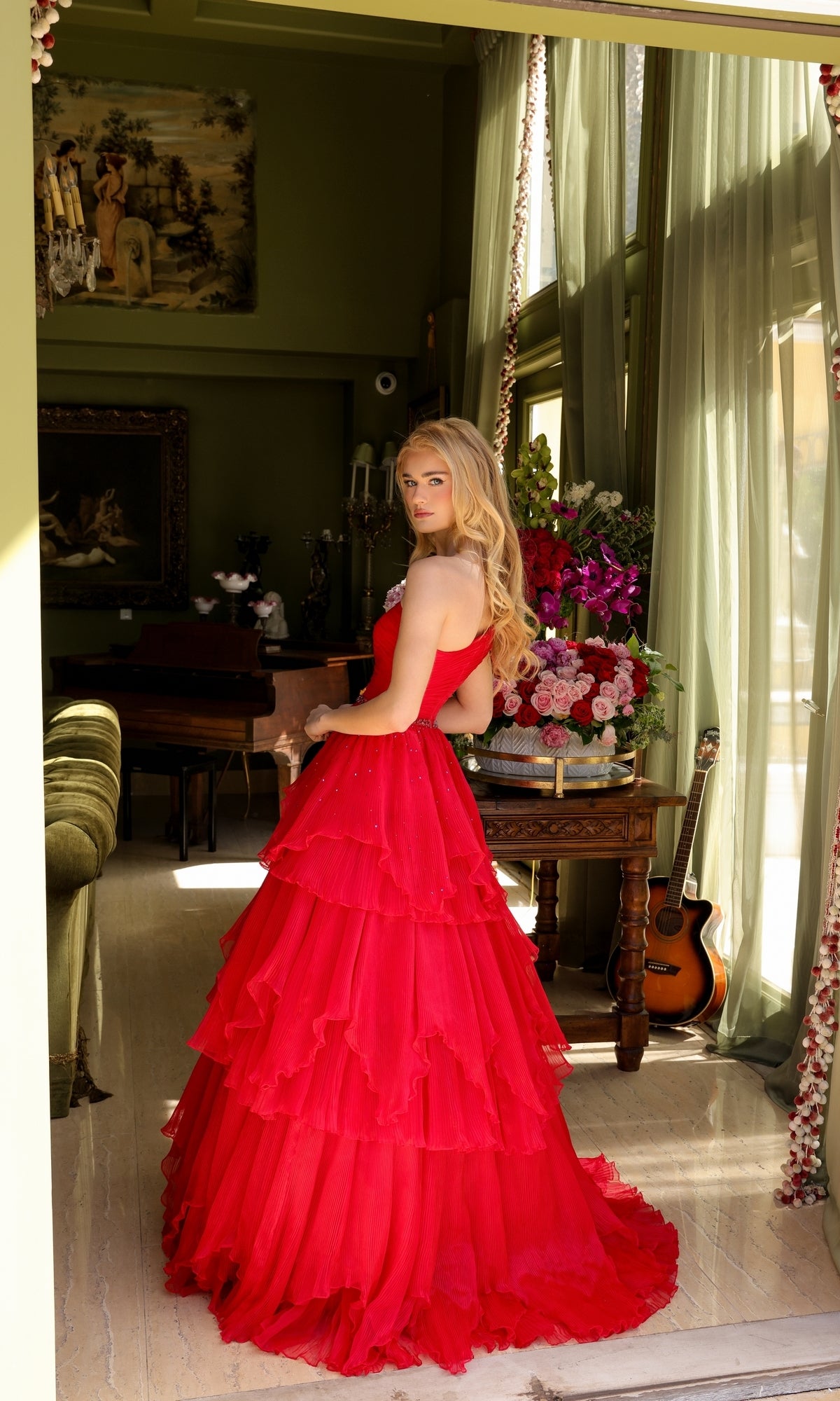 A woman stands indoors near a window, wearing the Ava Presley 29131 one-shoulder tiered prom gown, with flowers, a table, and a guitar visible in the background.