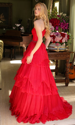 A woman with long blonde hair wears the Ava Presley 29131 One-Shoulder Tiered Prom Gown, standing indoors near a table adorned with a large bouquet of flowers and a guitar.