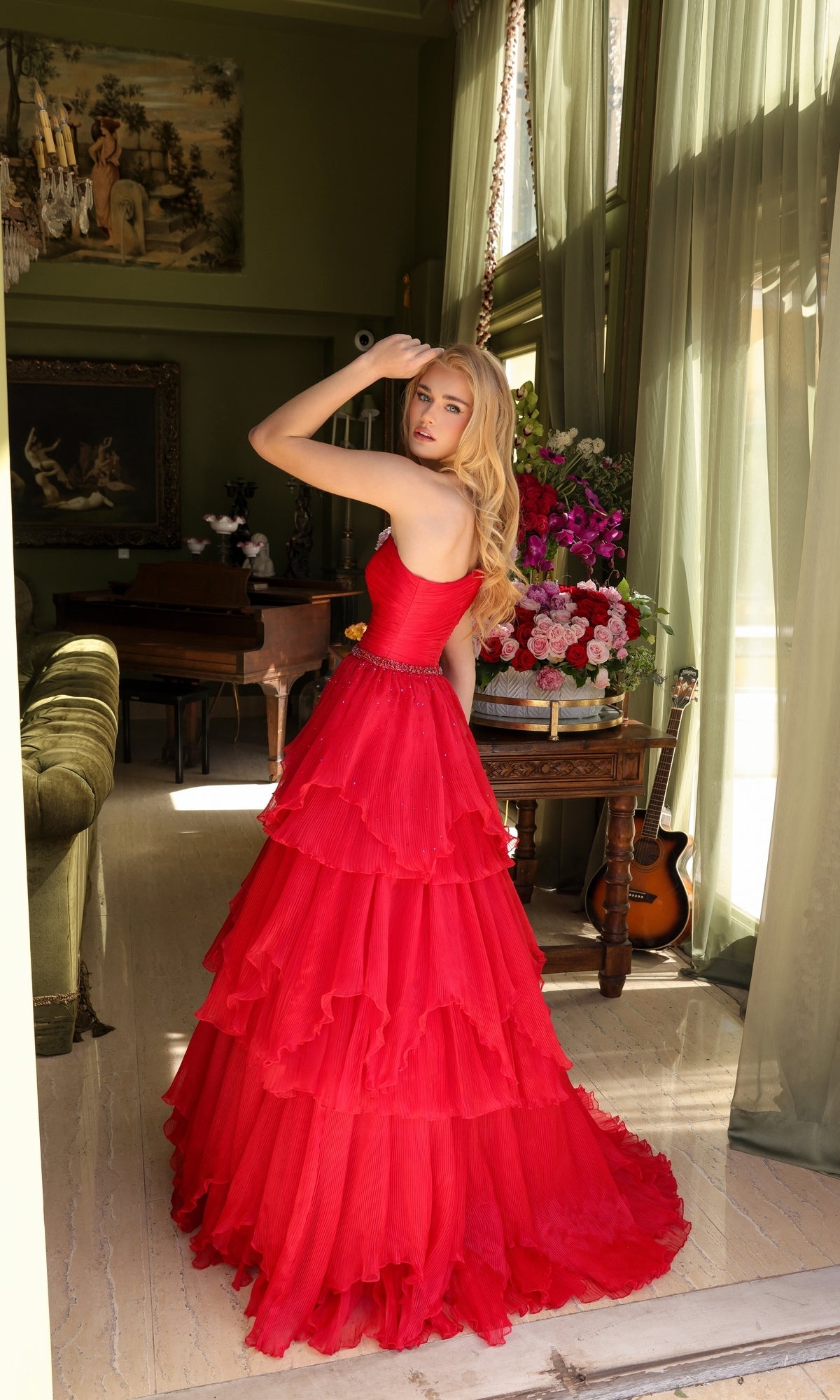 A woman wears the Ava Presley 29131 One-Shoulder Tiered Prom Gown indoors, facing backward and glancing over her shoulder, with flowers, a guitar, and large windows in the background.