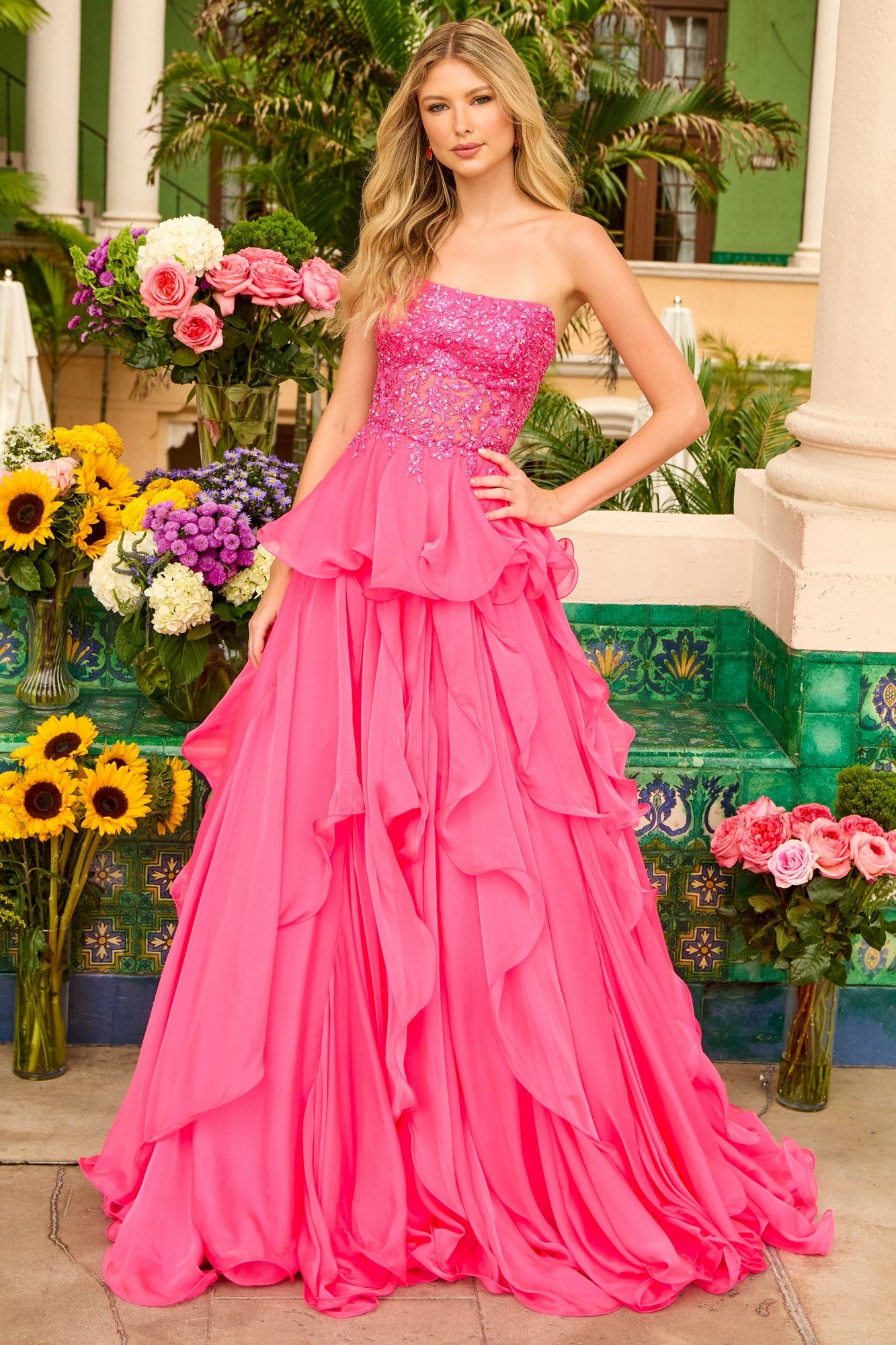 A woman stands outdoors in the Ava Presley 29133 strapless ruffle ball gown, featuring a bright pink layered skirt and embroidered corset bodice, surrounded by colorful flowers and lush greenery.